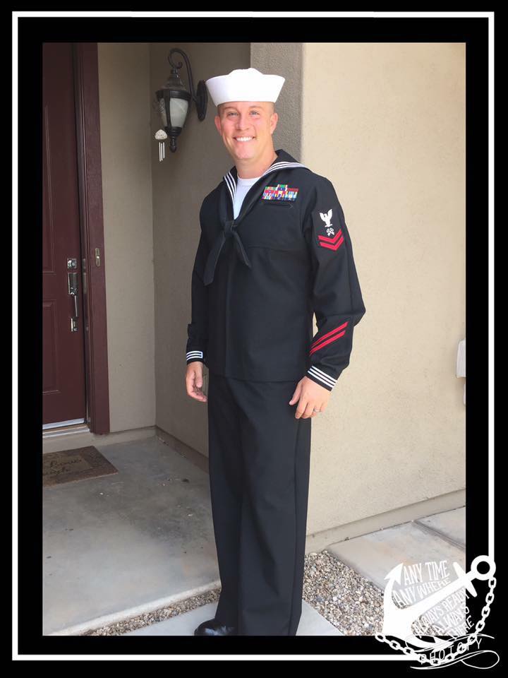 Adam Scott in a navy blue uniform and white sailor cap standing in front of a building entrance.