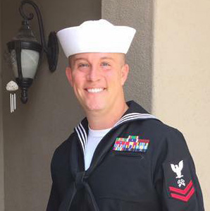 Adam Scott in U.S. Navy uniform smiling and standing in front of a light brown wall.