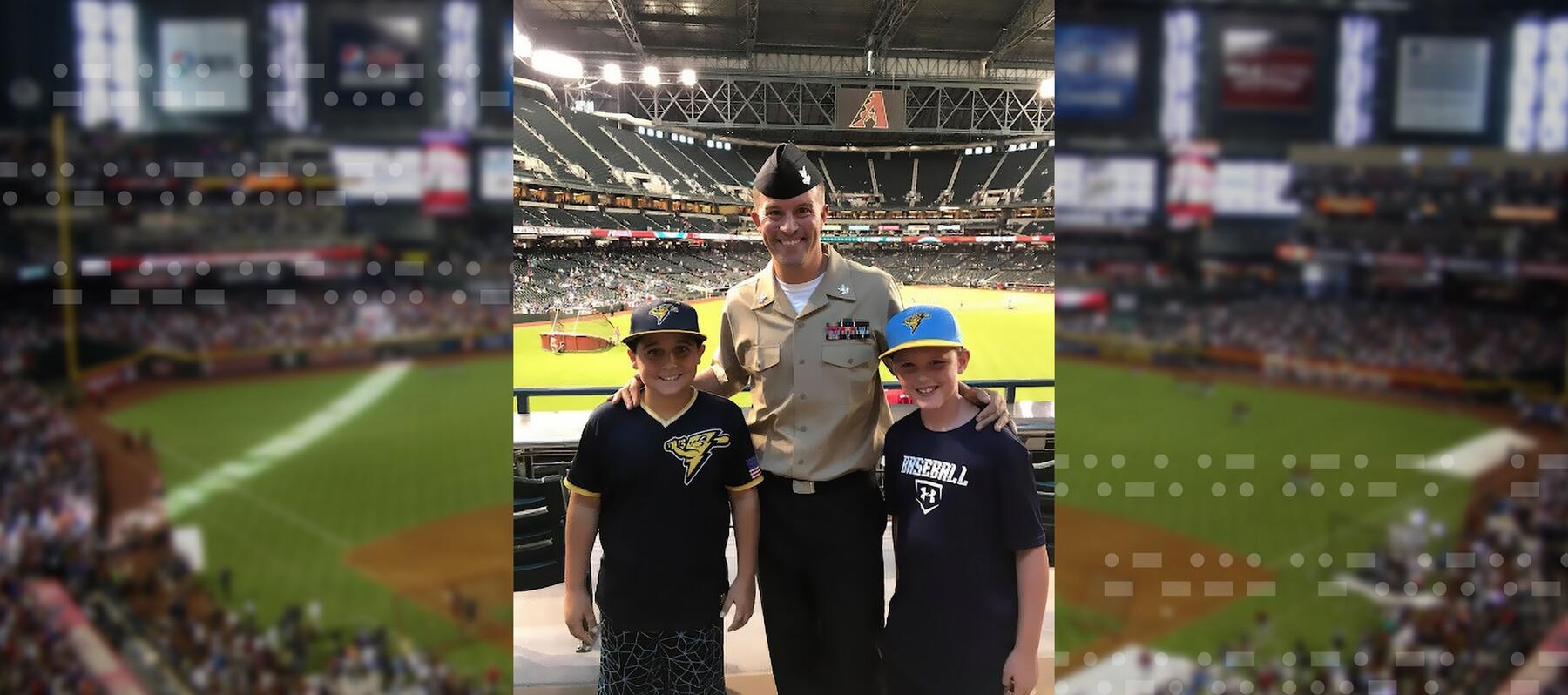 A person in a military uniform stands between two kids in a baseball stadium.